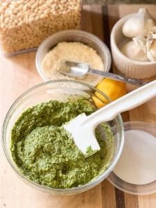 Portrait shot of finished pesto sauce featuring springtime dandelion greens. Grated parmesan, a zested lemon, and whole heads of garlic can be seenin the upper right corner of the photo.