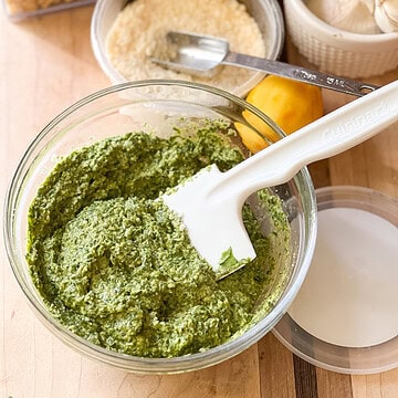 Portrait shot of finished pesto sauce featuring springtime dandelion greens. Grated parmesan, a zested lemon, and whole heads of garlic can be seenin the upper right corner of the photo.