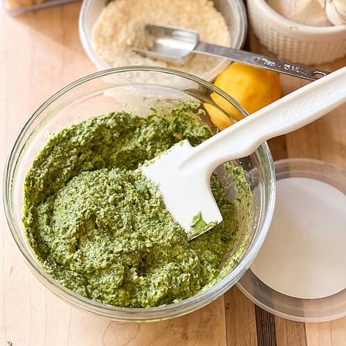 Portrait shot of finished pesto sauce featuring springtime dandelion greens. Grated parmesan, a zested lemon, and whole heads of garlic can be seenin the upper right corner of the photo.