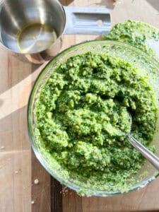 Flatlay image of prepared Kale Pesto with Toasted Walnuts. A silver spoon holds a small scoop of the green pesto flaked with grated parmesan.