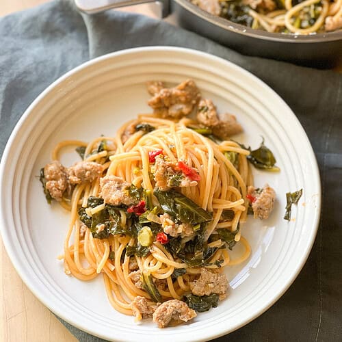 Portrait photo of a white ceramic shallow bowl filled with cooked spaghetti tossed with dino rabe, broccoli greens, Italian sausage in a sauce of Calabrian chiles and tomato paste for a super simple and delicious Single Skillet Spaghetti.