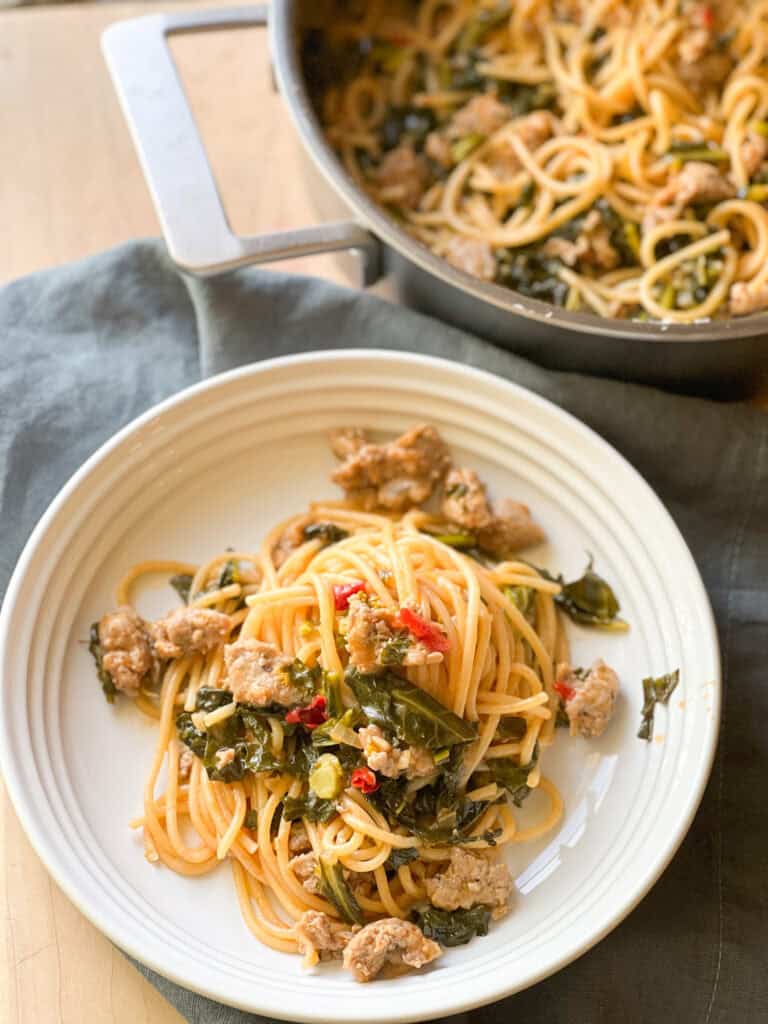 Portrait photo of a white ceramic shallow bowl filled with cooked spaghetti tossed with dino rabe, broccoli greens, Italian sausage in a sauce of Calabrian chiles and tomato paste for a super simple and delicious Single Skillet Spaghetti. 