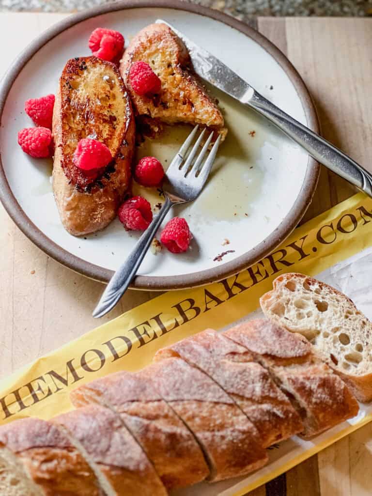 Flatlay photo of a dish in the upper left corner with two slices of baguette french toast recipe garnished with fresh berries and maple syrup. A fork and butter knife are sitting atop the plate with a piece of the french toast cut.