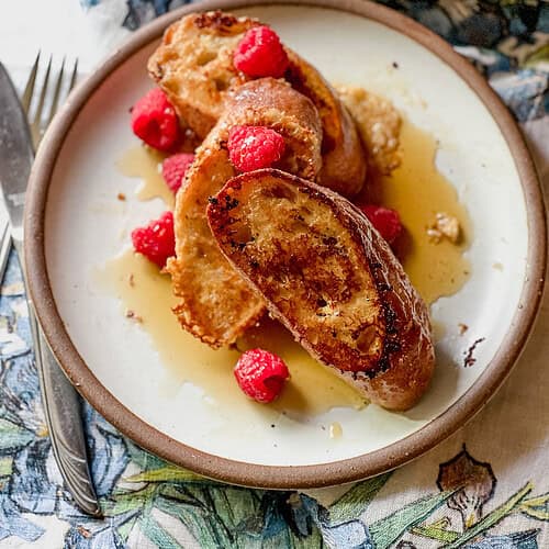 Portrait photo of finished dish of Baguette French Toast with Mascarpone Custard Recipe. The dish is garnished with fresh raspberries and maple syrup.