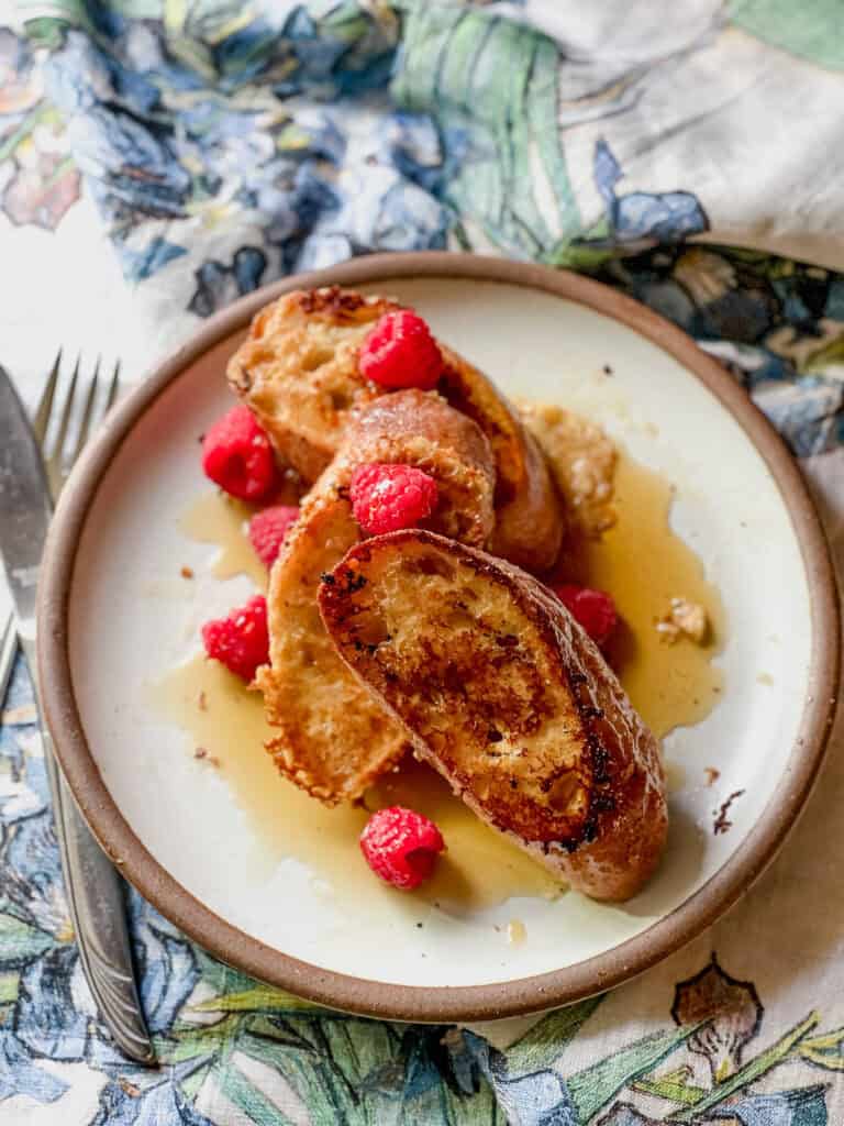 Portrait photo of finished dish of Baguette French Toast with Mascarpone Custard Recipe. The dish is garnished with fresh raspberries and maple syrup.