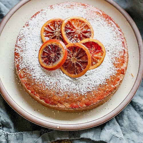 Portrait photo of the finished Blood Orange and Vanilla Olive Oil Snack Cake. It's garnished with powdered sugar and candied blood orange slices. The entire 9 inch cake round sits atop a cream colored ceramic plate on top of a sage colored linen napkin.