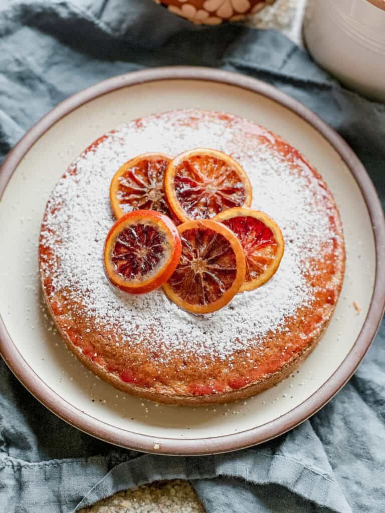 Portrait photo of the finished Blood Orange and Vanilla Olive Oil Snack Cake. It's garnished with powdered sugar and candied blood orange slices. The entire 9 inch cake round sits atop a cream colored ceramic plate on top of a sage colored linen napkin.