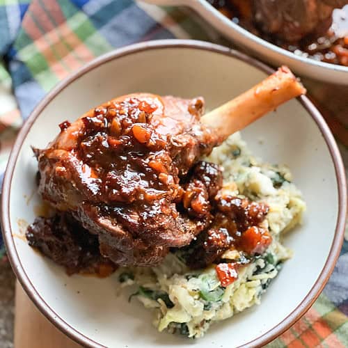 Flatlay shot of the finished dish for Fall-Apart Tender Braised Lamb Shanks with Red Wine & Herbs plated over a bed of brown butter colcannon.