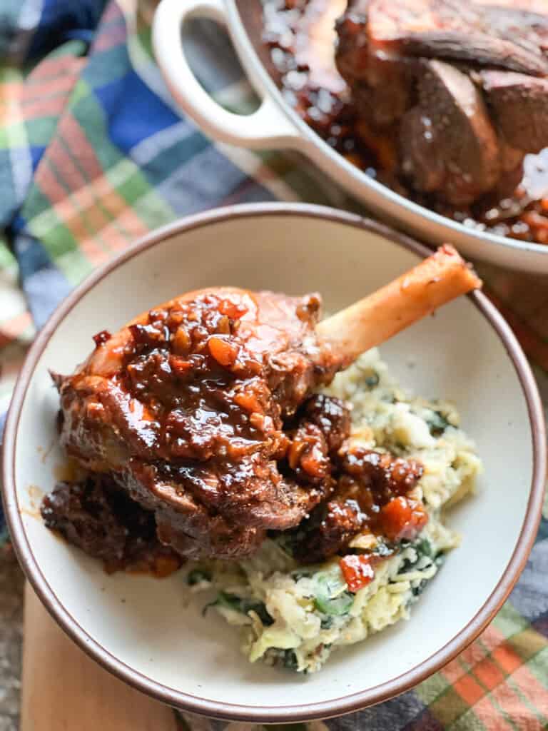 Flatlay shot of the finished dish for Fall-Apart Tender Braised Lamb Shanks with Red Wine & Herbs plated over a bed of brown butter colcannon.