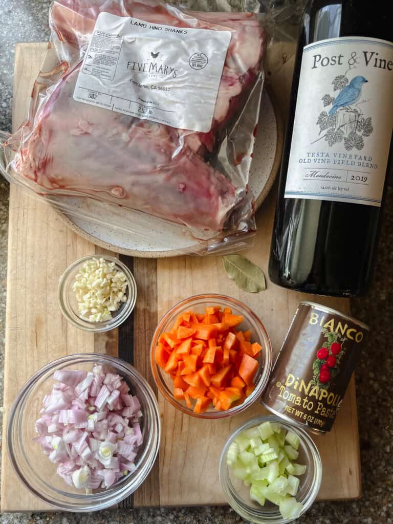 Ingredient layout photo for the Fall-Apart Braised Lamb Shanks with Red Wine and Herbs. Featuring a bottle of Napa Valley red wine, local lamb shanks, diced carrots, shallot, celery, minced garlic, bay leaf, and tomato paste.