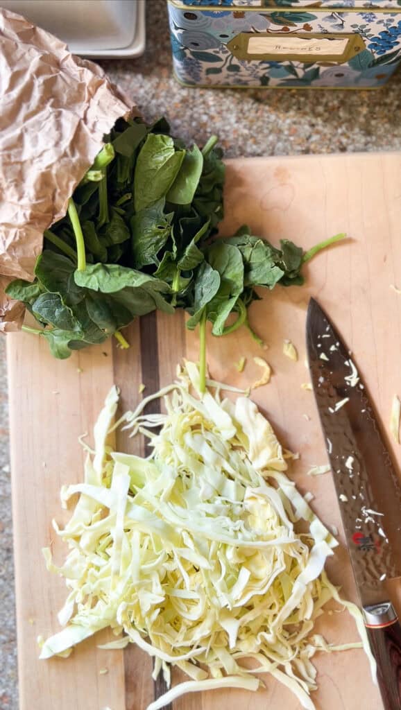 Flatlay photo of chopped spinach and cabbage in meal prep mode for the Brown Butter Colcannon with Cabbage, Spinach, and Leeks. The greens are displayed chopped on top of a wooden cutting board. A chef's knife is displayed to the bottom right corner of the photo.