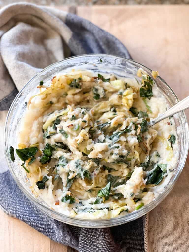 Portrait of the finished side dish: Brown Butter Colcannon with Cabbage, Spinach, and Leeks. Served in a large bowl accented with a kitchen towel.