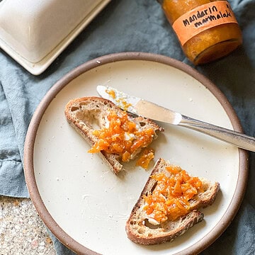 Flatlay image of a halved piece of toast with a pat of butter and a dollop of Mandarin Marmalade spread across the top of the toast. The jar of marmalade labeled "Mandarin Marmalade" is visible in the upper right corner of the photo.