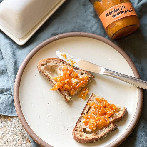 Flatlay image of a halved piece of toast with a pat of butter and a dollop of Mandarin Marmalade spread across the top of the toast. The jar of marmalade labeled "Mandarin Marmalade" is visible in the upper right corner of the photo.