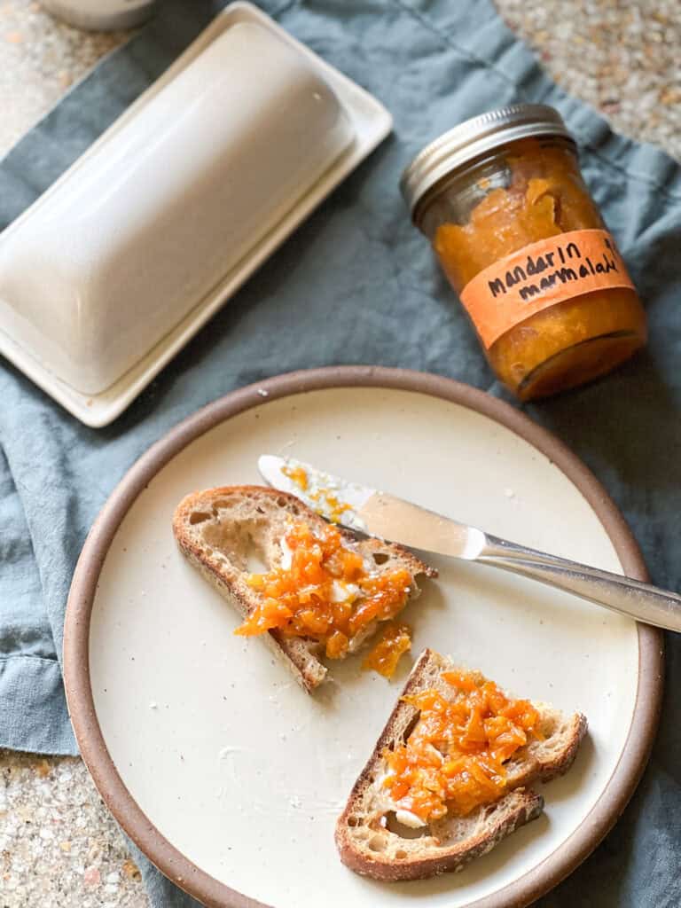 Flatlay image of a halved piece of toast with a pat of butter and a dollop of Mandarin Marmalade spread across the top of the toast. The jar of marmalade labeled "Mandarin Marmalade" is visible in the upper right corner of the photo.