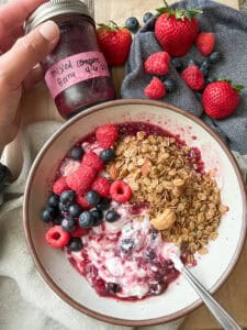 Flatlay image of a shallow white ceramic bowl filled with plain Greek yogurt with swirls of the Mixed Berry and Raspberry Compote Recipe. The compote is topped with homemade granola and fresh local berries.