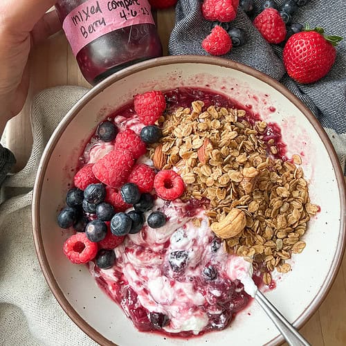 Flatlay image of a shallow white ceramic bowl filled with plain Greek yogurt with swirls of the Mixed Berry and Raspberry Compote Recipe. The compote is topped with homemade granola and fresh local berries.