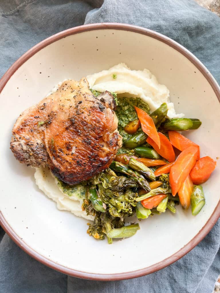 Finished dish of roast herb chicken thighs on a bed of white bean-potato mash served with the roasted carrots and asparagus recipe with a drizzle of seasonal pesto.