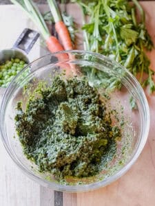 Flatlay photo of prepared Carrot Top, Radish Top, and Parsley Recipe. The finished pesto is accented with fresh carrots and remaining carrot tops towards the upper right portion of photo.
