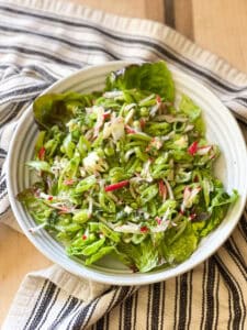 Flatlay photo of the assembled Easy Sugar Snap Peas, Radish, and Mint Salad Recipe. A white ceramic shallow bowl cradles a single serving portion of the salad. A white and blue striped kitchen towel is underneath the bowl atop a wooden cutting board.