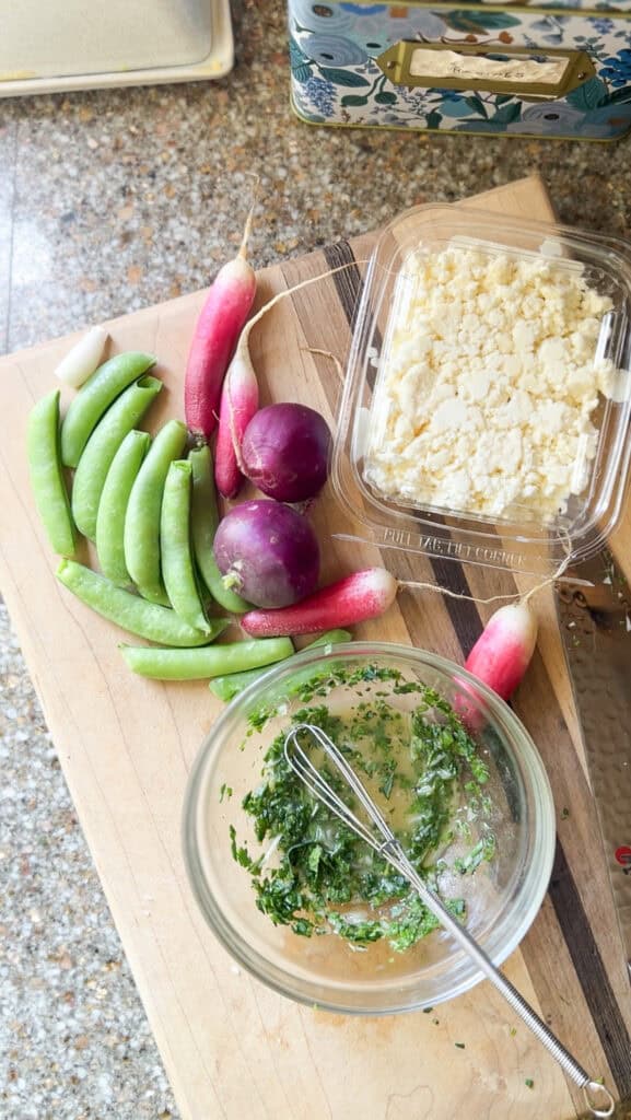 Flatlay of initial ingredients for the Easy Sugar Snap Peas, Radish, and Mint Salad Recipe. Trimmed sugar snaps, 4 French Breakfast radishes, a tub of feta, and a shallow bowl containing salad dressing ingredients are seen sitting atop a wooden cutting board.
