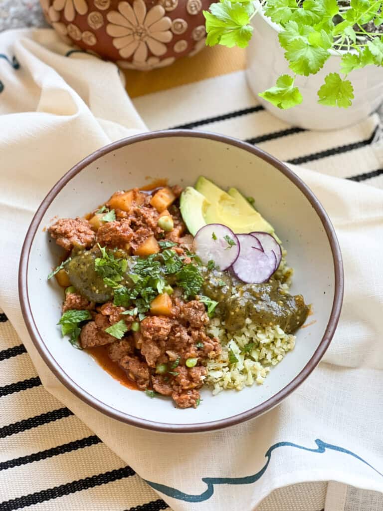 Flatlay photo of the finish dish of Grandma's Picadillo Recipe made with ground lamb and seasonal veggies. Garnished with slices of radish, avocado, and chopped fresh cilantro and mint.