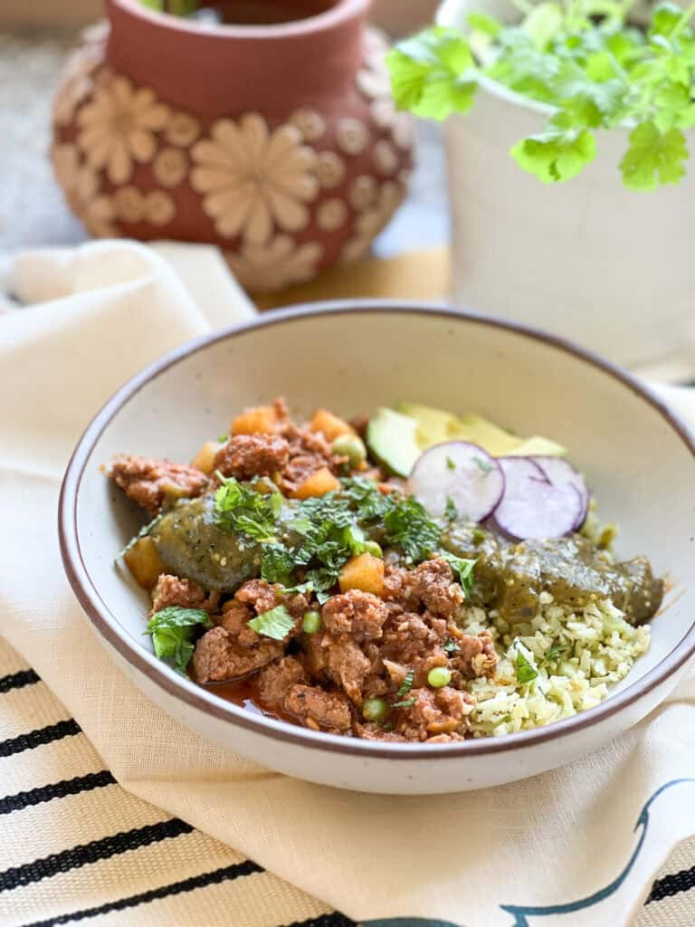 Flatlay photo of the finish dish of Grandma's Picadillo Recipe made with ground lamb and seasonal veggies. Garnished with slices of radish, avocado, and chopped fresh cilantro and mint.