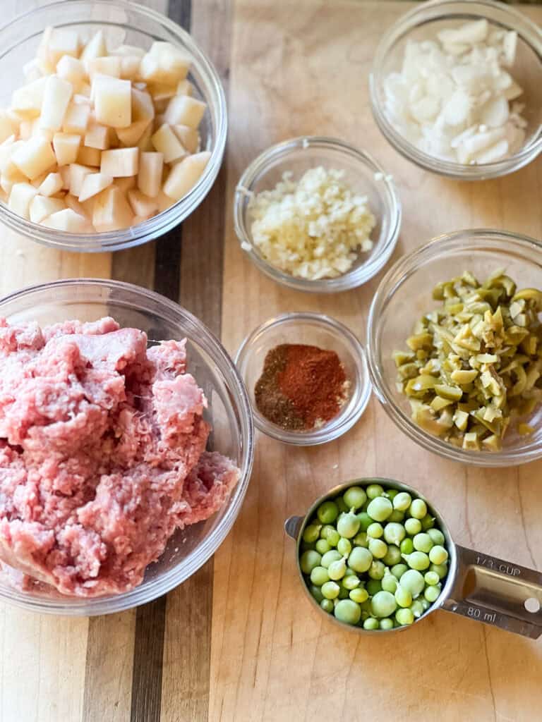 Flatlay photo of ingredients laid out for Grandma's Picadillo Recipe featuring ground lamb and seasonal vegetables like English peas and new potatoes. Clockwise from top left are diced new potatoes, diced shallots, minced garlic, chopped green olives, shelled English peas, assorted spices and seasonings, and ground lamb.