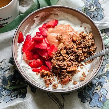 Portrait photo of a bowl of yogurt topped with Rhubarb Curd, fresh strawberries, and homemade granola.
