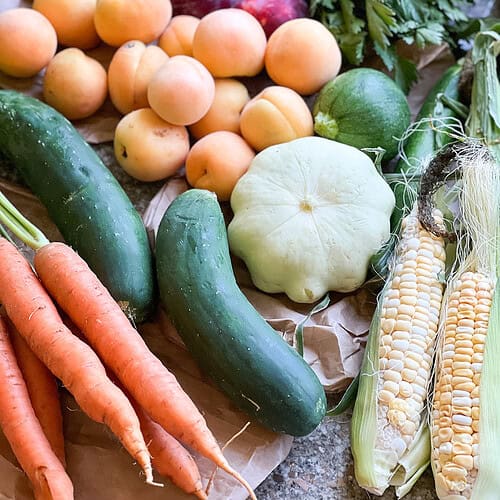 Flatlay photo of summertime produce for Dinner Plans Week 28 featuring corn, summer squash, cucumbers, carrots, apricots, plums, and parsley.