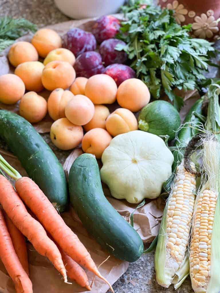 Flatlay photo of summertime produce for Dinner Plans Week 28 featuring corn, summer squash, cucumbers, carrots, apricots, plums, and parsley.