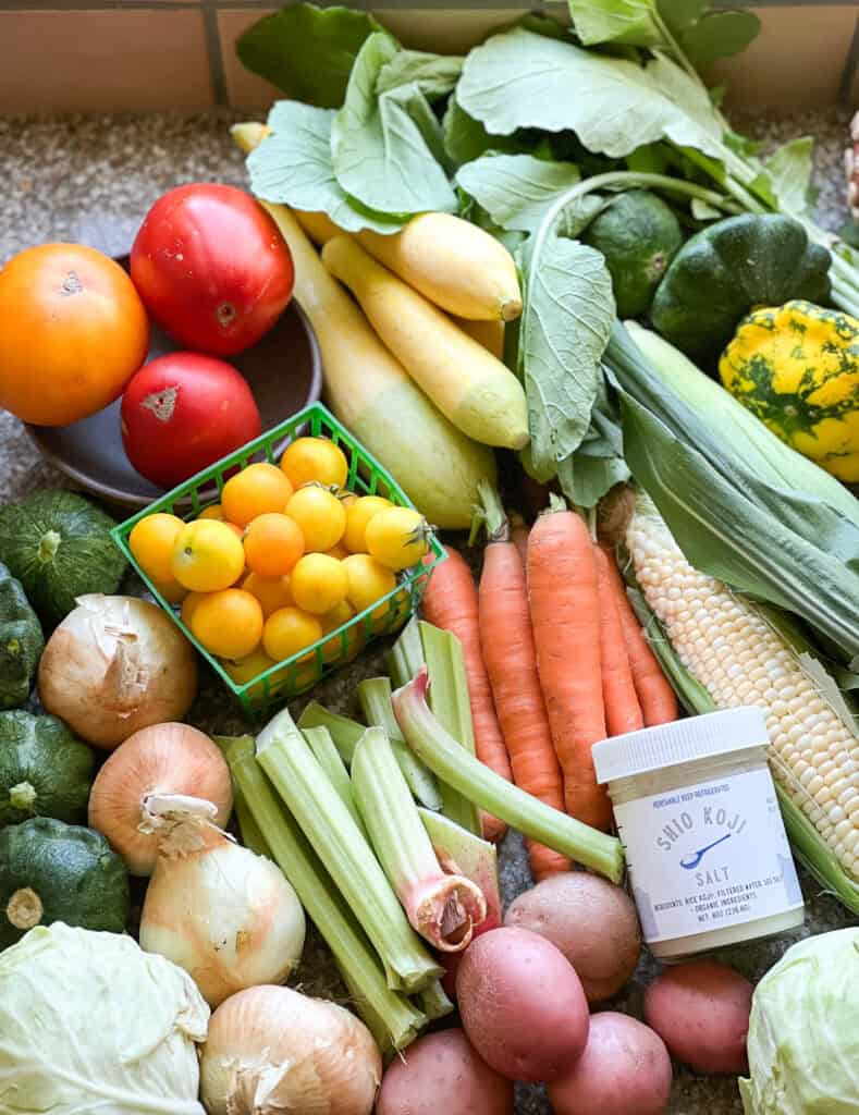Portrait photo of summer produce for Dinner Plans Week 30 featuring tomatoes, specialty summer squash, corn, carrots, rhubarb,red potatoes, walla walla onions, cabbage, and daikon radish. 