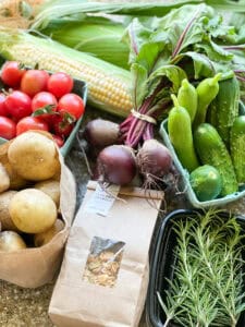 Portrait photo of summertime produce for Dinner Plans Week 31 featuring farm fresh corn, cherry tomatoes, potatoes, small batch granola, baby beets, fresh rosemary, and pickling cucumbers.