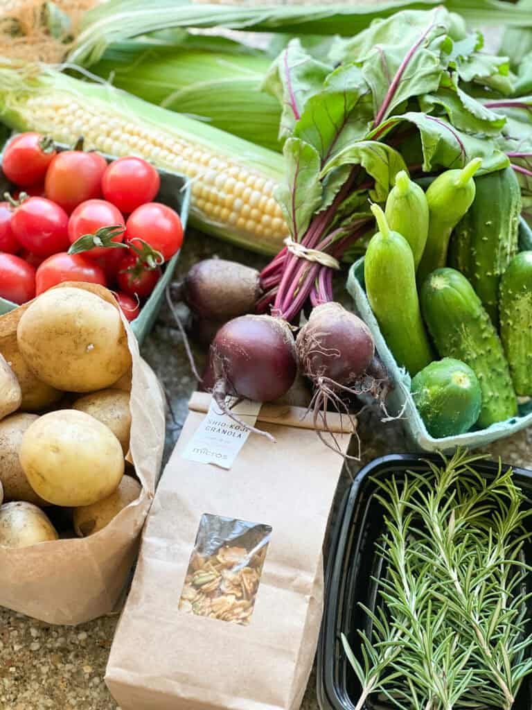 Portrait photo of summertime produce for Dinner Plans Week 31 featuring farm fresh corn, cherry tomatoes, potatoes, small batch granola, baby beets, fresh rosemary, and pickling cucumbers. 