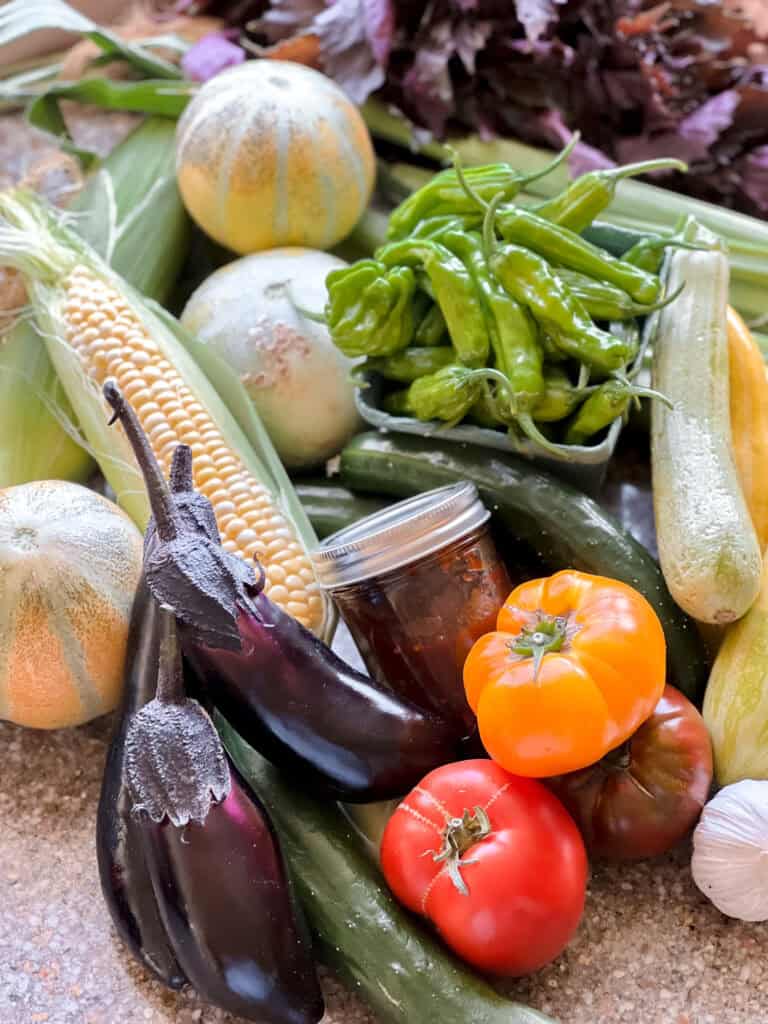 Flatlay photo of summer produce for Dinner Plans Week 33 featuring Japanese Eggplant, Heirloom Tomatoes, Shishito Peppers, Garlic, Summer Squash, Sweet Corn, Shiso, and melon. 