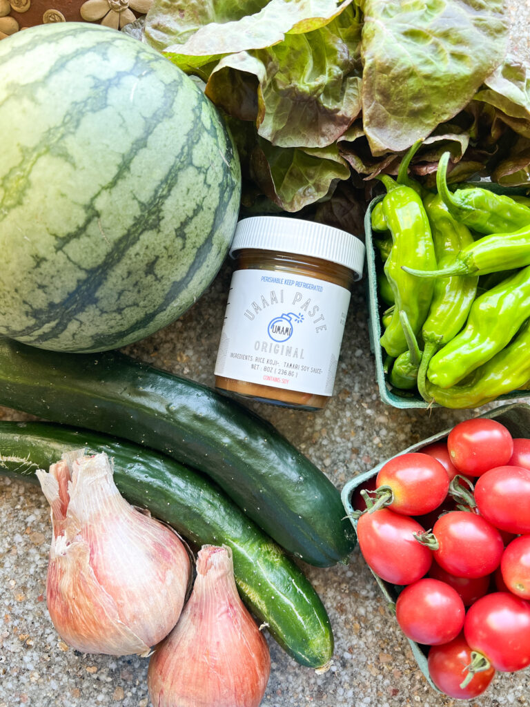 Flatlay photo of late summer produce featuring watermelon, Sunpeach cherry tomatoes, shallots, cucumbers, shishito peppers, and green leaf lettuce.