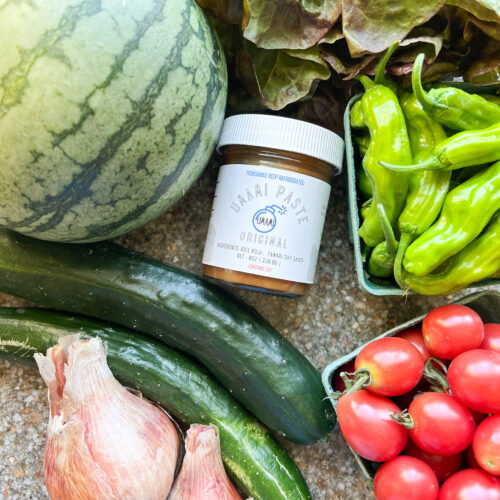 Flatlay photo of late summer produce featuring watermelon, Sunpeach cherry tomatoes, shallots, cucumbers, shishito peppers, and green leaf lettuce.