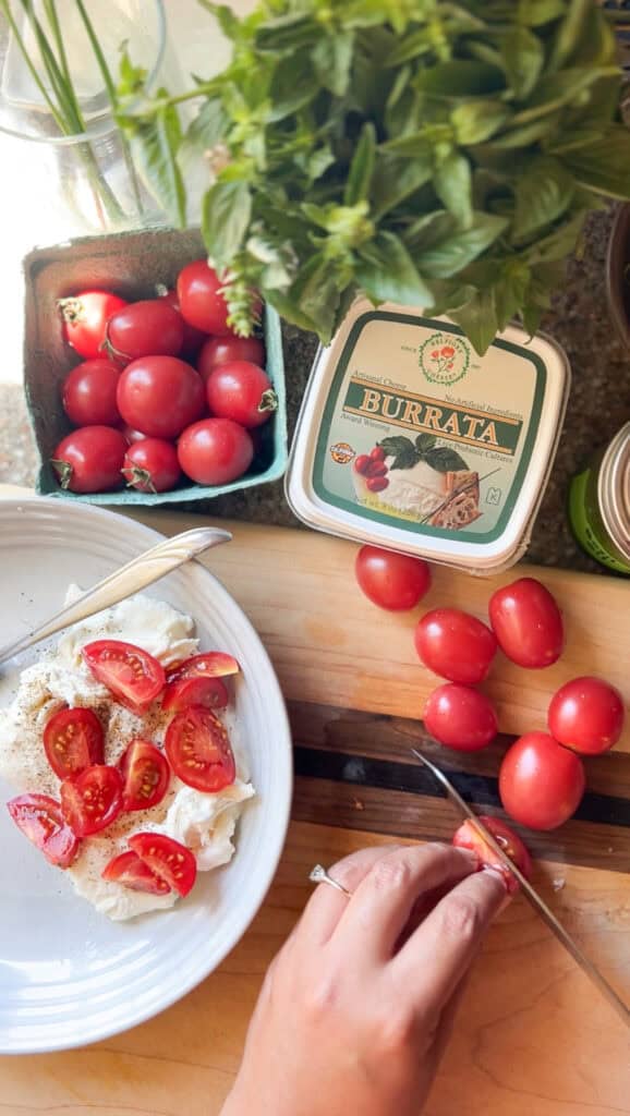 A brown woman's hand cuts cherry tomatoes for the Caprese Salad with Cherry Tomatoes and Basil Oil. Basil and tomatoes are shown in the upper portion of the screen. 