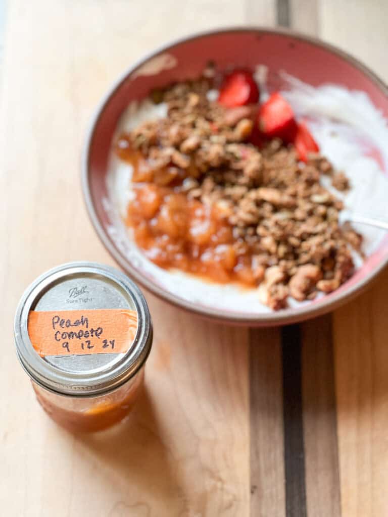 An offset jar of Peach Compote adjacent to the bowl of yogurt with swirls of Peach Compote and homemade granola.