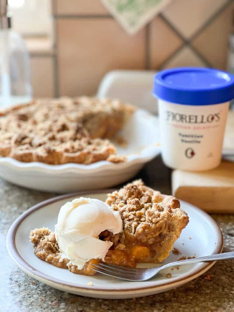 A portrait photo of a slice of Vanilla Bourbon Baked Peach Pie recipe with a scoop of vanilla gelato.