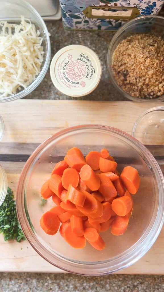 overhead photo of the ingredients assembled for this seasoned carrots recipe featuring creme fraiche, carrots, herbs, cheese, and breadcrumbs.