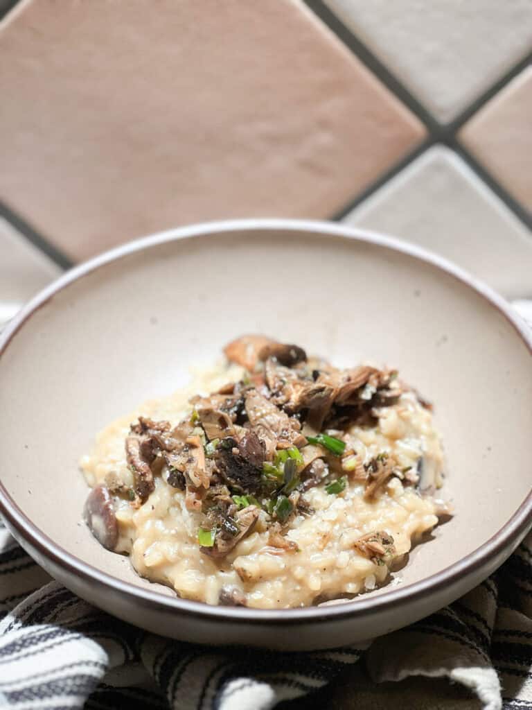 Portrait photo of mushroom risotto generously served in a shallow bowl topped with sauteed mushrooms.