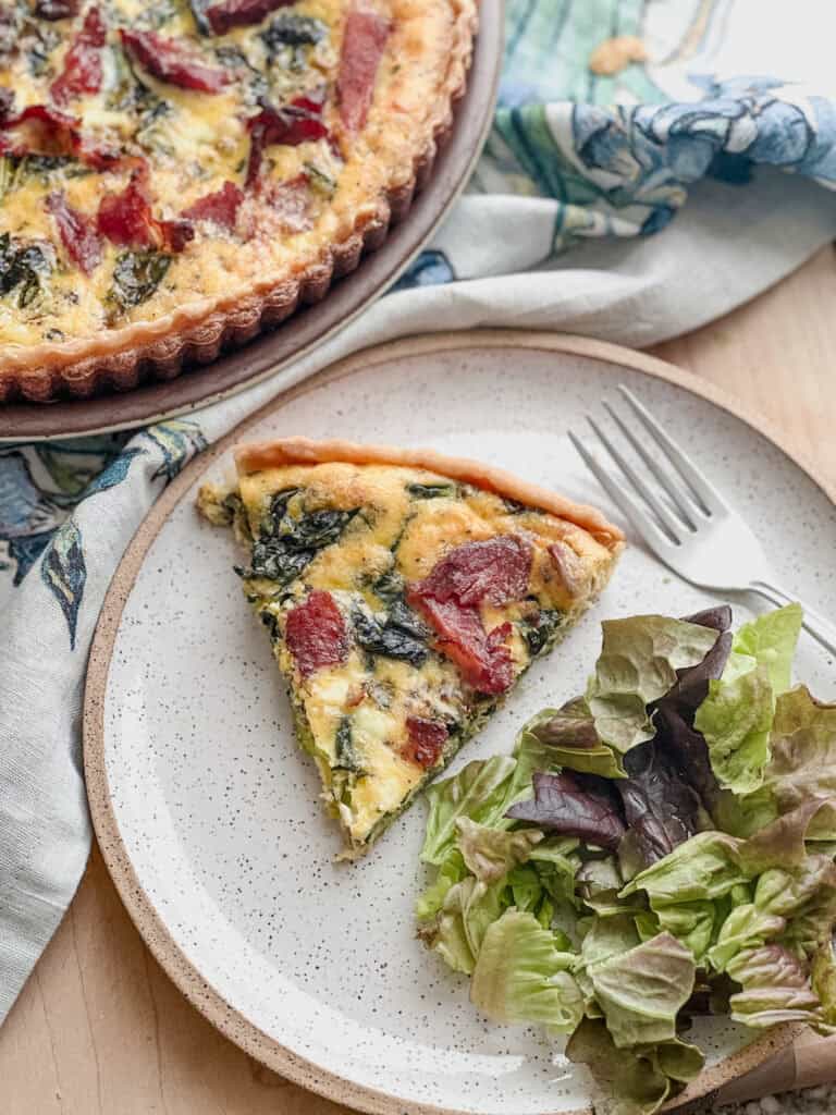 A slice of the Christmas Quiche Recipe sits atop a pie plate with a side serving of salad. 