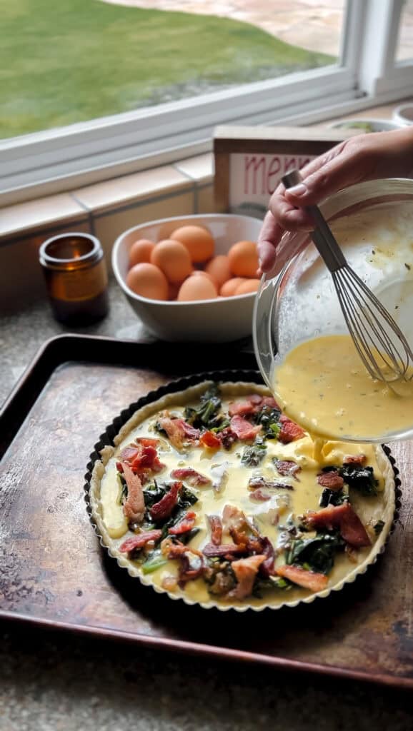 Portrait shot of a woman's hands holding a bowl of egg custard for the quiche filling. The custard is seen being poured over the prepared pie crust with layered filling. 