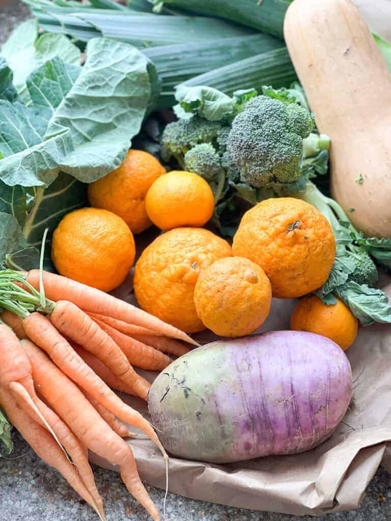 Portrait image of early spring produce featuring purple daikon radish, carrots, mandarins, broccoli, collard greens, butternut squash, and spinach.