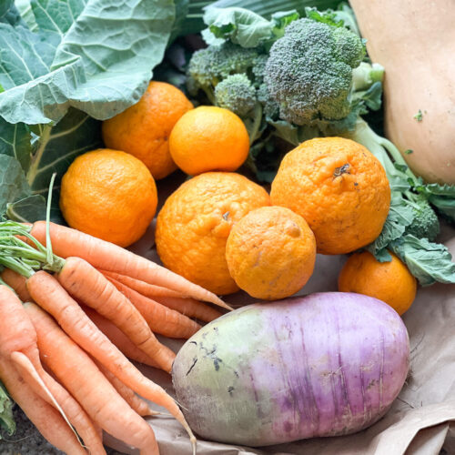 Portrait image of early spring produce featuring purple daikon radish, carrots, mandarins, broccoli, collard greens, butternut squash, and spinach.