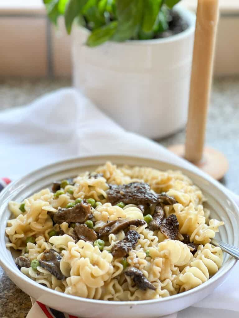 Portrait photo of the Pasta with Sauteed Morel Mushrooms and Peas Recipe. A white ceramic pasta bowl holds a generous portion of the pasta and sits atop a French-style linen kitchen towel.