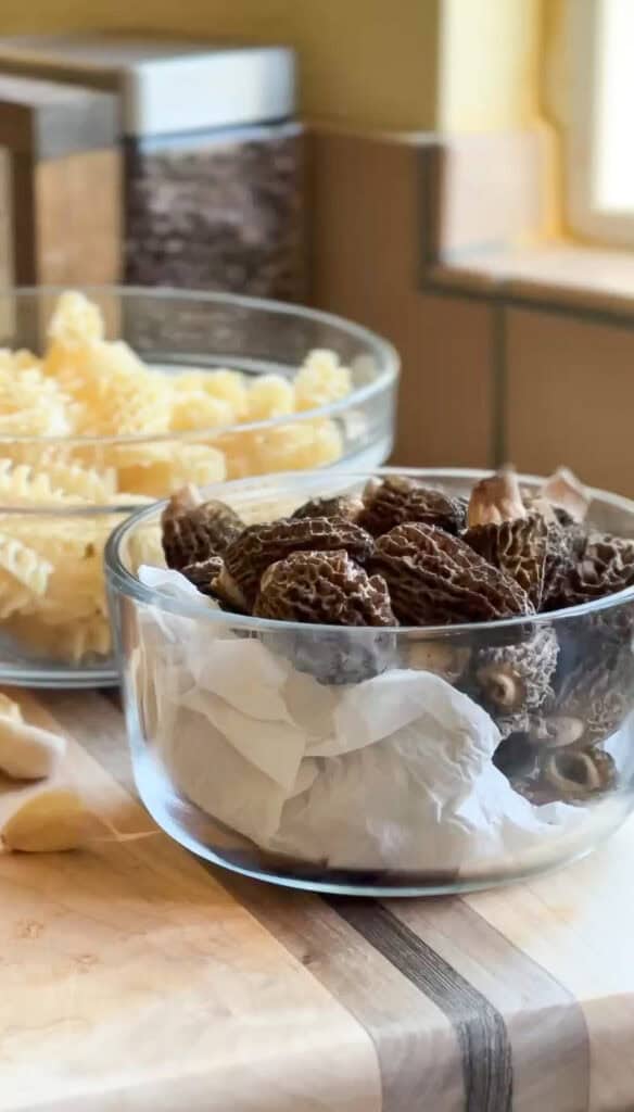 Portrait shot of cleaned morel mushrooms in front of a bowl of dried pasta in preparation for the easy weeknight mushroom pasta recipe.