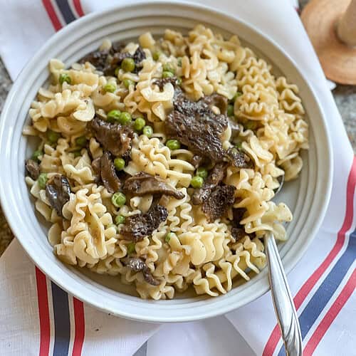 Portrait photo of the Pasta with Sauteed Morel Mushrooms and Peas Recipe. A white ceramic pasta bowl holds a generous portion of the pasta and sits atop a French-style linen kitchen towel.