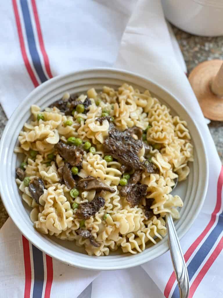 Portrait photo of the Pasta with Sauteed Morel Mushrooms and Peas Recipe. A white ceramic pasta bowl holds a generous portion of the pasta and sits atop a French-style linen kitchen towel. 